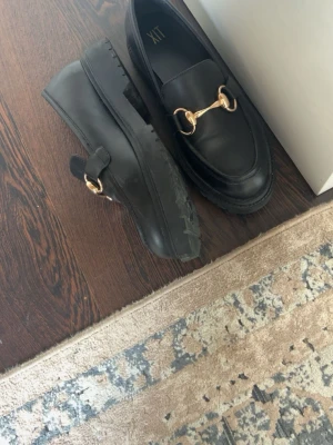 Svarta loafers med gulddetalj XIT - Stilrena svarta loafers från XIT med chunky sula och snygg guldfärgad metallspänne över foten. Skorna har en klassisk form och är tillverkade i slätt material som ger en clean look. Perfekta för dig som gillar trendiga och tidlösa skor.