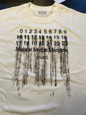 Maison Margiela vit t-shirt med tryck - Säljer en vit t-shirt från Maison Margiela med svart tryck framtill som har siffror och texten 'Maison Martin Margiela PARIS' i en cool drip-effekt. Klassisk rund hals och korta ärmar. Perfekt för dig som gillar streetstyle och designerplagg.