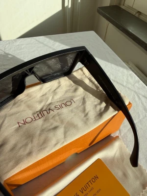 Svarta Louis Vuitton monogram-solglasögon - Säljer ett par svarta solglasögon från Louis Vuitton med rektangulär form och ikoniskt monogrammönster på glasen. Bågarna är i blank svart plast och glasen har en mör ? ton. Perfekt accessoar för dig som vill sticka ut med lyxig stil.