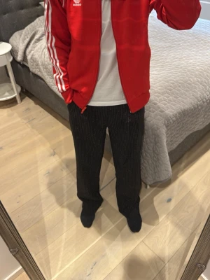 Röd Adidas ziptröja med vita ränder - Säljer en röd Adidas ziptröja med klassiska vita ränder längs ärmarna och Adidas-logga på bröstet. Tröjan har dragkedja framtill, ribbade muddar och är tillverkad i ett sportigt material. Perfekt för en sportig och avslappnad stil.