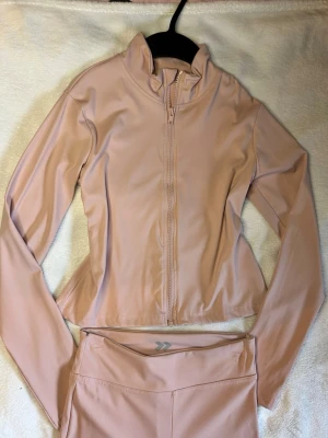 Ljusrosa träningsset med jacka och bootcut tights - Säljer ett snyggt träningsset i ljusrosa med långärmad zip-jacka och matchande bootcut tights. Setet är gjort i ett stretchigt och mjukt material som passar perfekt för gym eller löpning. Jackan har hög krage och dragkedja framtill. 149 kr för båda