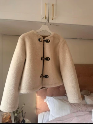 Beige teddyjacka med toggleknäppning - Säljer en beige teddyjacka i mjukt, fluffigt material med tre svarta toggleknappar framtill. Jackan har rund halsringning, raka ärmar och en något boxig, croppad passform. 