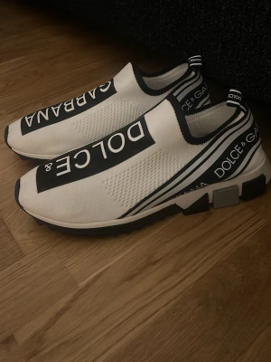 Dolce & Gabbana vita slip-on sneakers - Helt oanvända stilrena vita slip-on sneakers från Dolce & Gabbana med svarta detaljer och tydlig logga på ovansidan och hälen. Skorna har en sportig design med elastisk ovandel i textil och en chunky sula i gummi. Perfekta för dig som gillar streetwear och exklusiv stil. Kontakta gärna mig vid frågor eller för mer information!☺️