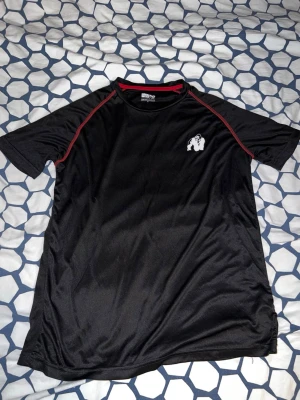 Svart tränings-tshirt Gorilla Wear - Svart tränings-tshirt från Gorilla Wear med röda detaljer längs axlarna och logotyp på bröstet och ryggen. Materialet är lätt och andas, perfekt för gym eller sport. Snygg passform och cool design med gorilla-motiv.