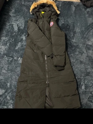 Svart lång dunjacka från Canada Goose - Säljer en svart lång dunjacka från Canada Goose med huva och avtagbar fuskpäls. Jackan har dragkedja framtill, diskret logga på ärmen och quiltad design. Perfekt för kalla vinterdagar och riktigt varm. Klassisk och stilren modell.