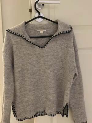 Grå stickad tröja med mörkblå kant - Mysig grå stickad tröja med mörkblå dekorativ kant längs krage, ärmslut och nederkant. Tröjan har en bred krage med V-ringning och avslappnad passform. Perfekt för lager på lager och enkel att styla till jeans eller kjol.