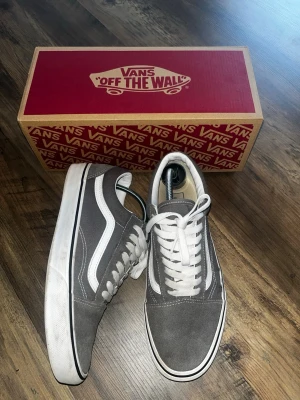 Gråa Vans Old Skool sneakers - Säljer ett par klassiska Vans Old Skool sneakers i grå mocka med vita detaljer och vit sula. Skorna har den ikoniska vita sidan, vita skosnören och låg profil. Perfekta för dig som gillar streetwear och enkel stil.