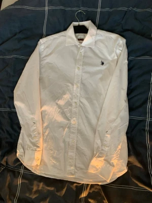 Vit skjorta från Polo Ralph Lauren - Klassisk vit skjorta från Polo Ralph Lauren med broderad logga på bröstet. Skjortan har lång ärm, knappar framtill och är tillverkad i bomull. Perfekt för en clean och stilren look.