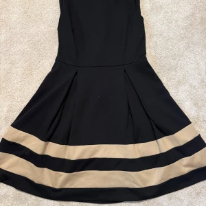 Svart och beige klänning Calvin Klein - Snygg svart klänning från Calvin Klein med beigea ränder nertill. Klänningen har en utsvängd kjol och markerad midja, vilket ger en klassisk och stilren siluett. Perfekt för dig som gillar enkel men elegant design.