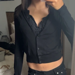 Svart cardigan med volangkant - Snygg svart cardigan med knappar framtill och volangkant vid halsringningen. Tröjan har lång ärm och ribbade muddar vid ärmslut och nederkant. Perfekt att styla till både jeans och kjol för en trendig look.