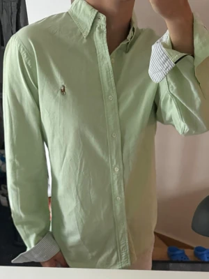Ljusgrön skjorta från Ralph Lauren - Säljer en ljusgrön skjorta från Ralph Lauren med klassisk broderad logga på bröstet. Skjortan har lång ärm, knappar framtill och button-down krage. Tillverkad i bomull och har randiga detaljer på insidan av manschetterna. Loggan har en liten tråddragning som man ser på sista bilden, Annars mycket bra skick 