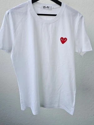 Vit t-shirt Comme des Garçons Play - Vit t-shirt från Comme des Garçons Play med det ikoniska röda hjärtat broderat på bröstet. Klassisk rund hals och korta ärmar. Tillverkad i mjuk bomull för en skön känsla och stilren look.