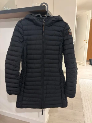 Svart dunjacka från Parajumpers - Snygg svart dunjacka från Parajumpers med huva och dragkedja framtill. Jackan är quiltad med smal passform och har logotyp på ena ärmen. Perfekt för kalla vinterdagar och håller dig varm med stil.