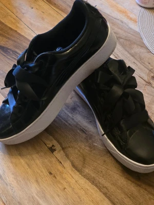 Svarta Puma sneakers med satinband - Stilrena svarta sneakers från Puma med glansig finish och breda satinliknande skosnören. Skorna har en tjock vit sula och klassisk låg profil. Perfekta för dig som vill ha en sportig men ändå trendig look. Har endast testat att ha dom på mig. 