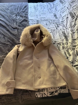 Beige kappa med päls från Gina Tricot - Stilren beige kappa från Gina Tricot med stor luva och mjuk fuskpäls runt kragen. Kappan har knäppning framtill, två fickor och dekorativ knapp vid ärmslut. Perfekt för kalla dagar och ger en snygg look till alla outfits.