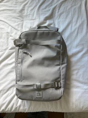 En bag - Snygg ljusgrå ryggsäck från Db med minimalistisk design. Ryggsäcken har justerbara axelband, en dragkedjeficka på framsidan och spännen för extra säkerhet. Tillverkad i slitstarkt material och har Db-logga både fram och bak.