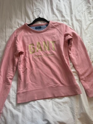 Rosa sweatshirt från GANT - Säljer en ljusrosa sweatshirt från GANT med guldfärgad text 'GANT New Haven' på bröstet. Tröjan har rund halsringning, långa ärmar och ribbade muddar. Perfekt för en avslappnad och trendig look. Storlek 152!