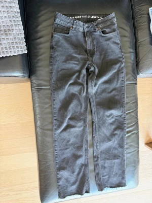 Grå mid wide jeans med raka ben - Snygga grå jeans med mid wide waist och raka ben. Klassisk femficksmodell med knapp och dragkedja framtill. Jeansen har en avslappnad passform och är perfekta för en trendig vardagslook. Längd 32, midja 27. Inget synligt märke.