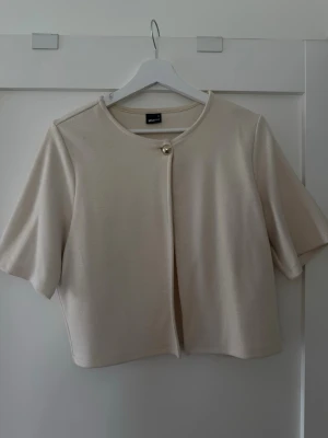 Kortärmad beige kofta med pärlknapp GinaTricot - Stilren beige kofta från GinaTricot med korta ärmar och rund halsringning. Koftan har en elegant pärlknapp framtill och är tillverkad i ett mjukt, tunt material som ger en clean look. Perfekt att slänga över en topp för en enkel men chic outfit.