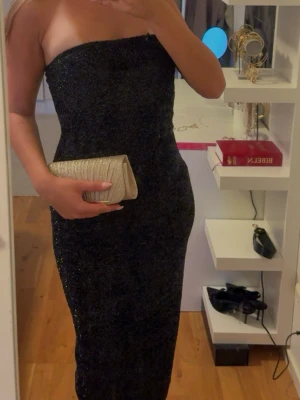 Svart glittrig axelbandslös klänning - Snygg svart axelbandslös klänning med glittrigt tyg som ger en festlig look. Klänningen är lång och har en tight passform som framhäver figuren (samt en liten slits baktill. 