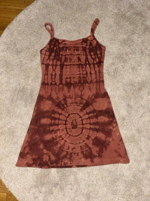 Brun batikmönstrad klänning - Säljer en kort, ärmlös klänning med smala axelband i brun batikstil. Klänningen har en figurnära passform och är tillverkad i ett mjukt, stretchigt material som känns bekvämt mot huden. Perfekt för dig som gillar unika mönster och en avslappnad look.