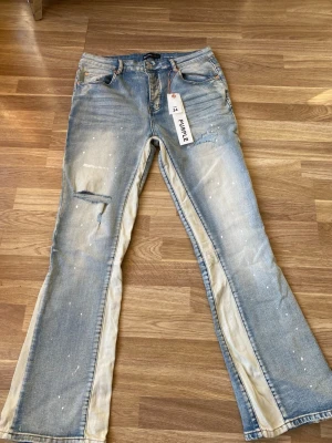 Ljusblå utsvängda jeans från Purple - Säljer ett par ljusblå jeans från Purple med utsvängda ben och beige paneler längs sidorna. Jeansen har slitningar, färgstänk och klassisk femficksdesign. Perfekta för dig som gillar en unik och trendig look.