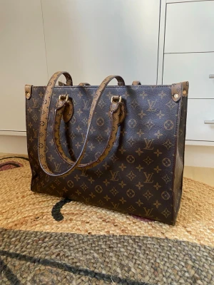  Toteväska - Stilren handväska från i klassiskt brunt monogrammönster med två långa handtag. Väskan är tillverkad i canvas med detaljer i läder och har rödrandigt innerfoder med tryckt logotyp. Perfekt för dig som gillar lyxiga accessoarer.