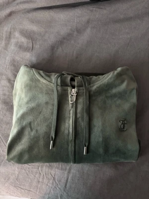 Grön hoodie från Juicy Couture - Mjuk grön hoodie från Juicy Couture i velourmaterial med dragkedja framtill och justerbar huva. Klassisk logga broderad på bröstet och snygga metalländar på snörena. 