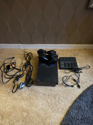 Sony PlayStation 2 med tillbehör - Säljer en svart PlayStation 2 konsol från Sony med två handkontroller, flera kablar och en multitap-adapter för fler spelare. Klassisk rektangulär design i plast med blå PS2-logga på ovansidan. Perfekt för dig som gillar retrospel och vill spela med vänner.