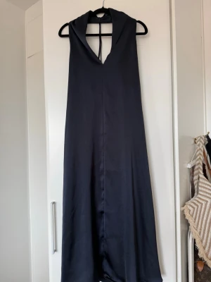 Marinblå långklänning med öppen rygg  - Elegant marinblå långklänning i ett glansigt material med djup v-ringning och öppen rygg som knyts i nacken. Klänningen har en rak och flowy passform, perfekt för dig som vill ha en stilren look med lite edge.