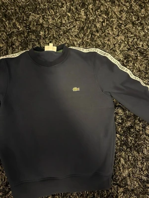 Mörkblå sweatshirt från Lacoste - Snygg mörkblå sweatshirt från Lacoste med klassisk passform. Tröjan har vita ränder med Lacoste-logga längs ärmarna och den ikoniska krokodilen broderad på bröstet. Perfekt för en sportig och stilren look.