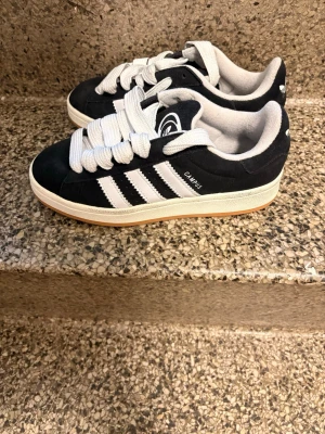Adidas Campus svarta sneakers - Svarta Adidas Campus sneakers med klassiska vita ränder på sidorna och vit logga på plösen. Ovandel i mocka, vit mellansula och gummisula. Breda vita skosnören och låg modell som ger en clean och tidlös vibe.