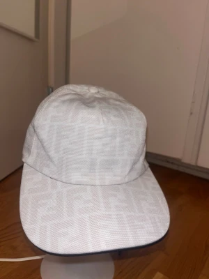 Vit keps med mönster - Snygg vit keps från med det klassiska FF-mönstret ton-i-ton över hela kepsen. Justerbar rem baktill och svart insida. Perfekt accessoar för att lyfta din streetwear-look.