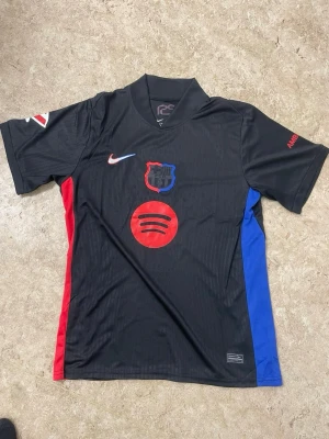 Svart FC Barcelona matchtröja Nike - Snygg svart FC Barcelona matchtröja från Nike med klubbmärke och Spotify-logga på bröstet. Tröjan har röda och blå detaljer på sidorna, korta ärmar och UNHCR-tryck på ryggen. Perfekt för dig som älskar fotboll och vill ha en unik supportertröja.