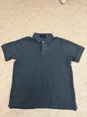 Mörkgrå pikétröja från Polo Ralph Lauren - Säljer en mörkgrå pikétröja från Polo Ralph Lauren med klassisk krage och knappar framtill. Tröjan har korta ärmar och ett broderat grönt Polo-logo på bröstet. Perfekt för dig som gillar stilrena och tidlösa plagg.