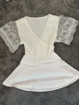 Vit baby doll  tröja  med spetsärmar - Söt vit babydoll  med omlott framtill och v-ringning. Tröjan har genomskinliga spetsärmar med blommigt mönster som ger en romantisk känsla. ”Kjolen” är lätt utsvängd och materialet känns mjukt och följsamt.