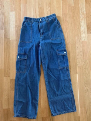 Blå cargobyxor med fickor - Snygga blå cargobyxor med bred passform och stora fickor på sidorna. Byxorna har resår i midjan och knäppning fram. Perfekta för en avslappnad och trendig look.