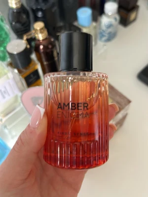 Amber Enigma parfym First Sense - Amber Enigma från First Sense är en parfym i en stilren glasflaska med orange till röd toning och svart cylinderformad kork. Flaskan har räfflad botten och elegant text på framsidan. Doften är inspirerad av varma, mystiska amber-noter. Använd vid ett tillfälle endast, i princip full flaska. 