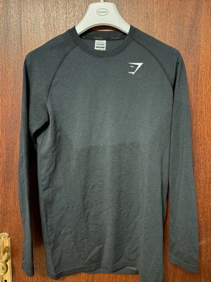 Svart långärmad träningströja Gymshark - Säljer en svart långärmad träningströja från Gymshark. Tröjan har diskret struktur i materialet, rund halsringning och vit Gymshark-logga på bröstet. Perfekt för gymmet eller löpning. Tillverkad i ett lätt och stretchigt funktionsmaterial.