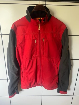 Röd och svart vindjacka från Fjällräven - Snygg vindjacka från Fjällräven i rött med svarta ärmar och detaljer. Jackan har hög krage, dragkedja framtill, två bröstfickor med dragkedja och två sidofickor. Tillverkad i slitstarkt material, perfekt för blåsiga dagar.