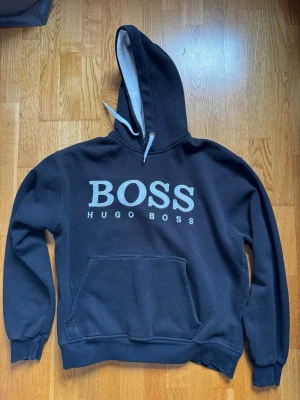 Svart hoodie från Hugo Boss - Snygg svart hoodie från Hugo Boss med stor vit logga på bröstet. Klassisk känguruficka framtill, ribbade muddar och justerbar huva med vita snören. Insidan av huvan är ljusgrå för en schysst kontrast. Perfekt för en chill och stilren look.