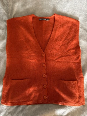 Orange stickad väst med knappar - Snygg orange stickad väst från Gudrun Sjödén med v-ringning och fem knappar framtill. Västen har två fickor fram och perfekt att slänga över en skjorta eller t-shirt för en cool lager-på-lager-look.