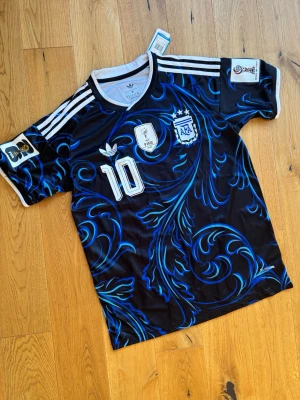 Argentina Messi matchtröja svart/blå - Säljer en svart och blå Argentina matchtröja från Adidas med Messi och nummer 10 på ryggen. Tröjan har vita detaljer, tre ränder på axlarna och ett unikt blått mönster över hela tyget. Tillverkad i lätt och ventilerande material, perfekt för fotboll eller träning. Helt oanvänd 
