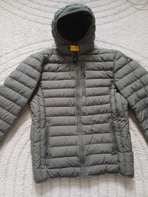 Grå dunjacka från Parajumpers - Snygg grå dunjacka från Parajumpers med huva och två dragkedjeförsedda fickor framtill. Jackan har det klassiska PJS-märket på ärmen och gul detalj vid halsen. Perfekt för kalla vinterdagar och riktigt lätt att bära. 12 till 14 åringar. Använd 2 till 3 gånger.