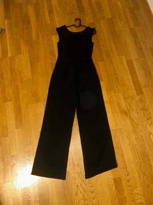 Svart ärmlös jumpsuit med v-ringning - Snygg svart jumpsuit med breda ben och ärmlös design. Djup v-ringning både fram och bak ger en stilren och modern look. Tillverkad i ett mjukt, följsamt tyg som sitter bekvämt. Perfekt för dig som vill ha en enkel men trendig outfit.