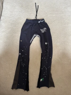 Svarta leggings från Gallery Dept. - Unika svarta leggings från Gallery Dept. med vitt tryck på höften och coola färgstänk över hela benen. Modellen har utsvängda ben med råa kanter och dragsko i midjan. Perfekta för dig som vill sticka ut med en edgy och kreativ stil.