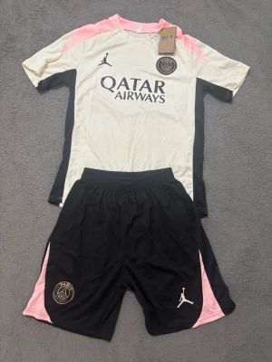 PSG träningsset vit/svart/rosa - Snyggt PSG träningsset med vit t-shirt och svarta shorts. Tröjan har rosa och svarta detaljer, PSG och Jordan-logga samt Qatar Airways-tryck. Shortsen är svarta med rosa inslag och PSG/Jordan-logga. Perfekt för fotboll eller träning.