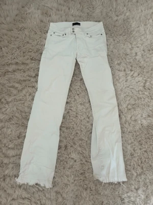 Vita jeans med råa kanter från Nudie - Säljer ett par vita jeans från Nudie med raka ben och råa, fransiga benslut. Jeansen har klassisk femficksmodell och orange broderi på bakfickorna. Perfekta för en avslappnad och trendig look.