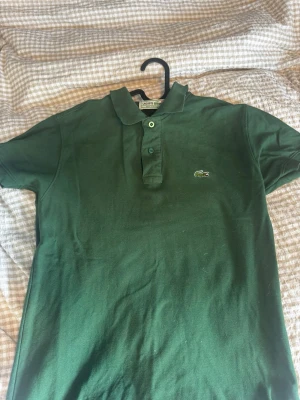 Grön pikétröja från Lacoste - Klassisk grön pikétröja från Lacoste med korta ärmar och den ikoniska krokodilloggan på bröstet. Tröjan har krage och knappar framtill. Perfekt till en avslappnad men ändå stilren look.