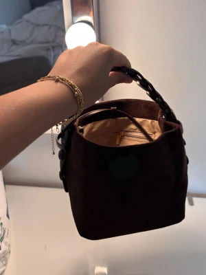Brun handväska - 🤎 Helt ny brun handväska 🤎  Säljer denna fina och trendiga väska i mörkbrun mockaliknande finish. Väskan är helt oanvänd och i nyskick då jag fick den i present, men den kom tyvärr aldrig till användning.  ✨ Elegant design med dekorativt handtag ✨ Avtagbart och justerbart axelband medföljer ✨ Rymlig nog för dina vardagsmåsten ✨ Perfekt till både vardag och fest  Inga skador eller anmärkningar – helt ny.  💌 Svarar snabbt på meddelanden och skickar snabbt efter köp. Hör gärna av dig vid frågor. 